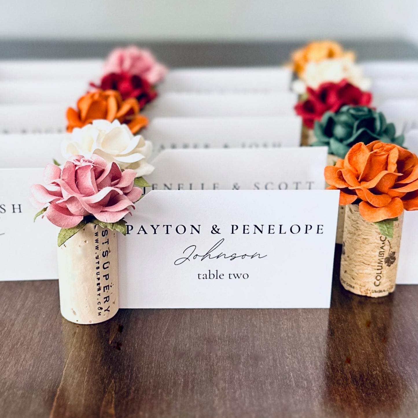 Decorative wine cork place card holders with floral elements on a wooden table. The custom color flowers are autumn wedding palette and include emerald, rust, terracotta, mauve, burgundy and white.