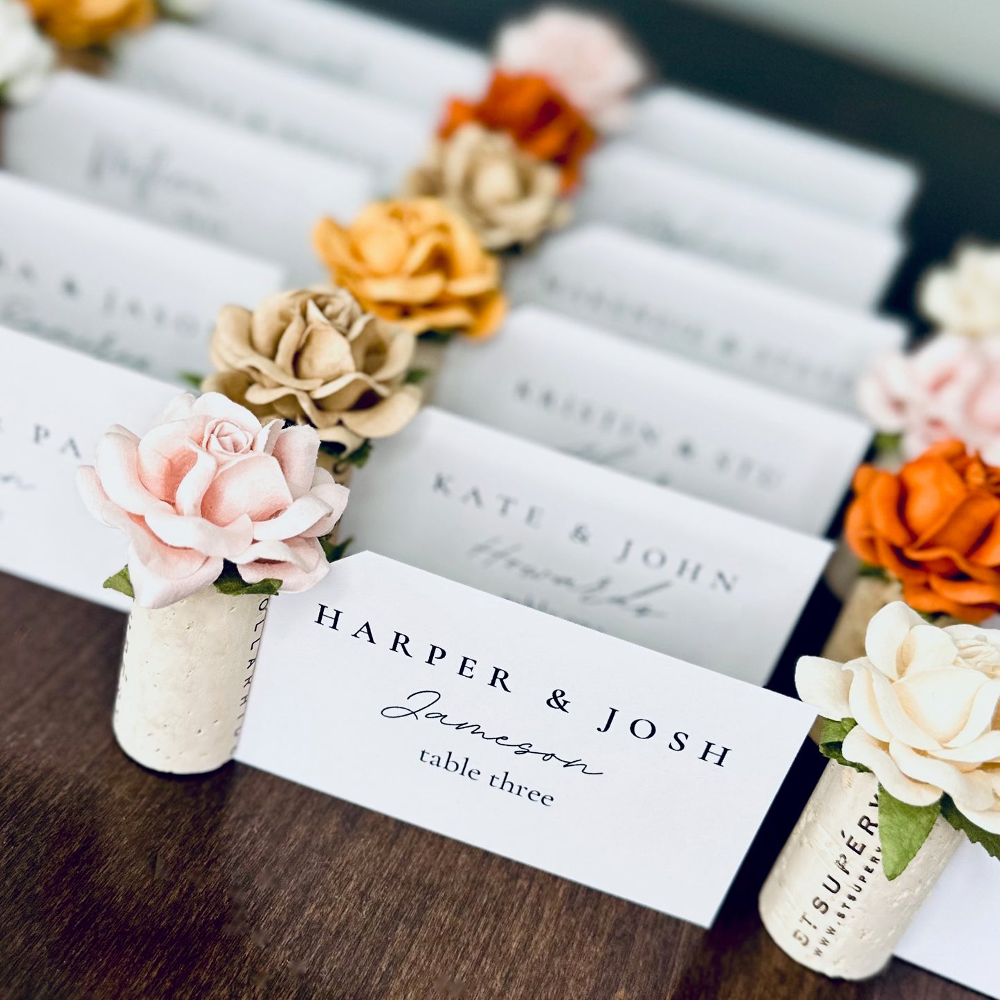 Guest place cards are displayed in vertical standing wine corks, with custom color roses on top. The colors suggest a fall wedding, in shades of rust, terracotta, gold, white, and blush pink. They are displayed on a wooden table.
