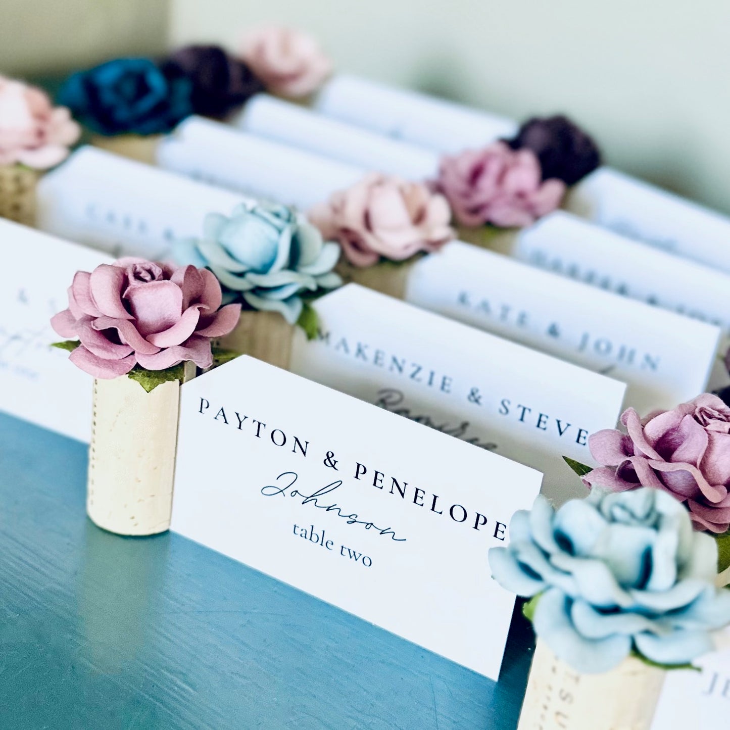 Printed guest name cards and table number assignments are displayed in wine cork place card holders with custom color rose embellishments. The roses are in shades of cornflower blue, wisteria, lavender, navy, and blush pink. Suggests a wedding place card table display.