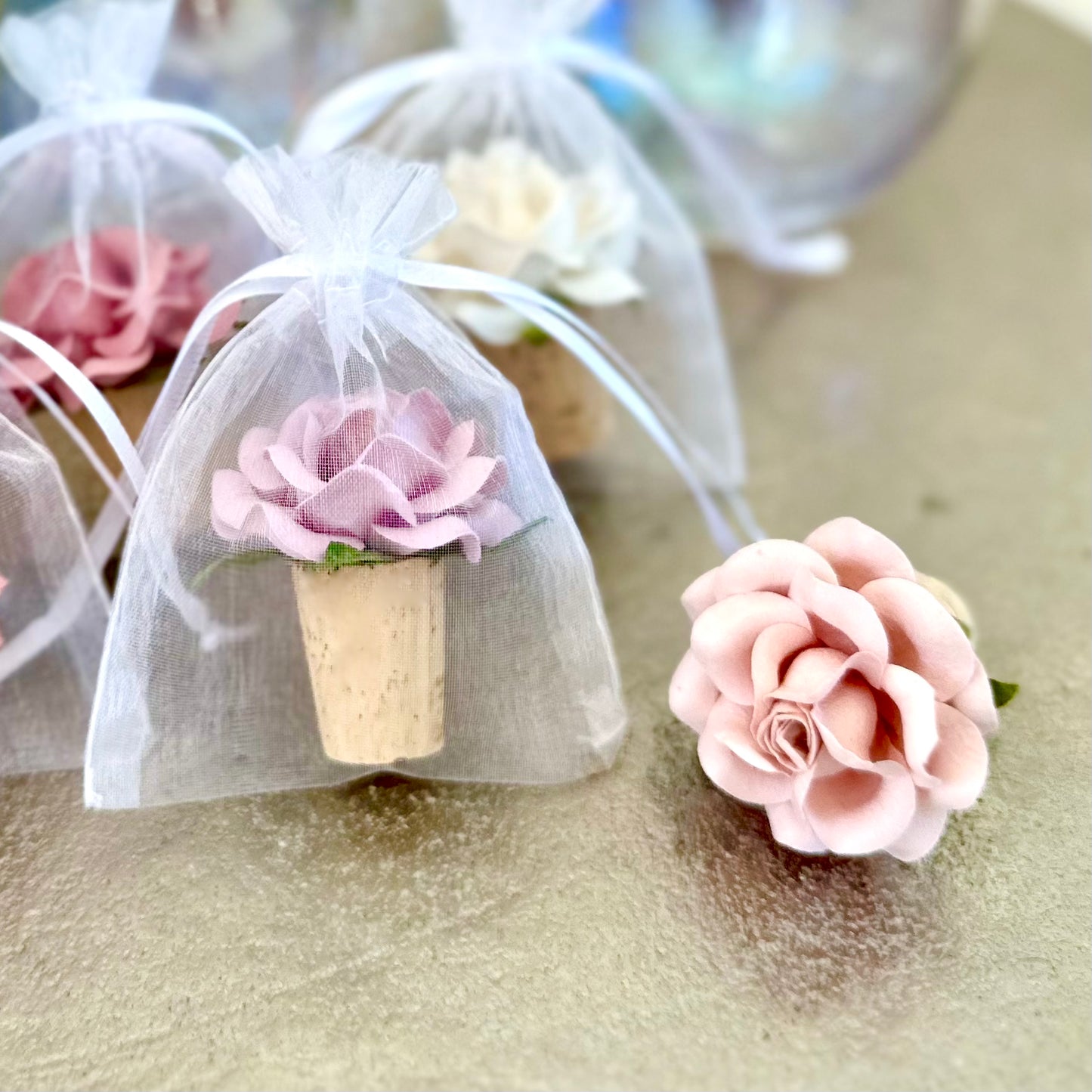 two wine bottle stopper wedding favors are displayed on a silver tabletop. There are beautiful paper flower blooms on top of the stoppers, one in lavender, one in Blush pink. The lavender wine bottle stopper is inside a white organza favor bag.