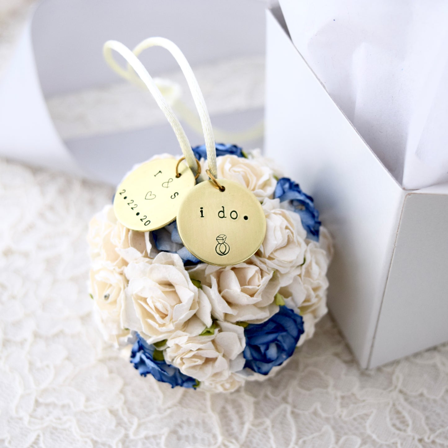 a beautiful blue & white paper flower wedding ornament with two personalized charms rests by a glossy white gift box, on a white lace surface.