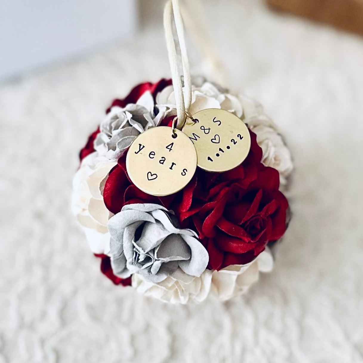 Floral 4th wedding anniversary ornament with red, burgundy, white, and gray flowers and personalized gold charms on an ivory lace surface.