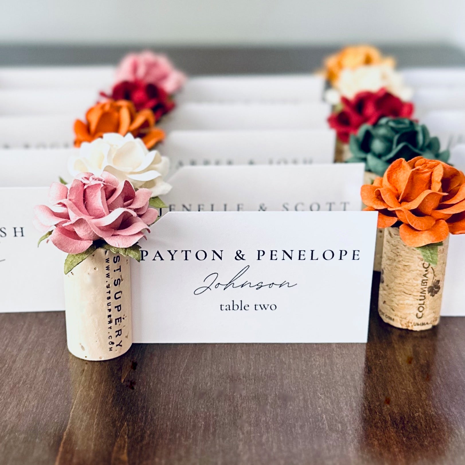 Decorative wine cork place card holders with floral elements on a wooden table.  The custom color flowers are autumn wedding palette and include emerald, rust, terracotta, mauve, burgundy and white.