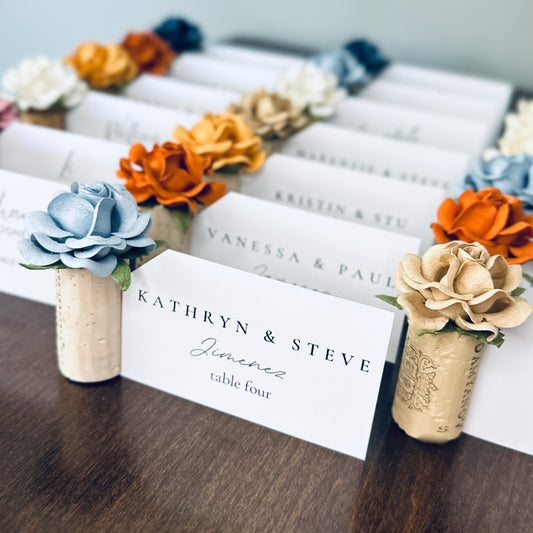 Wine cork place card holders with custom color roses in a fall wedding color palette.  Colors include rust, terracotta, tan, cornflower, navy blue, and white.  Guest name cards with calligraphy style font are displayed in the holders, with table assignments.