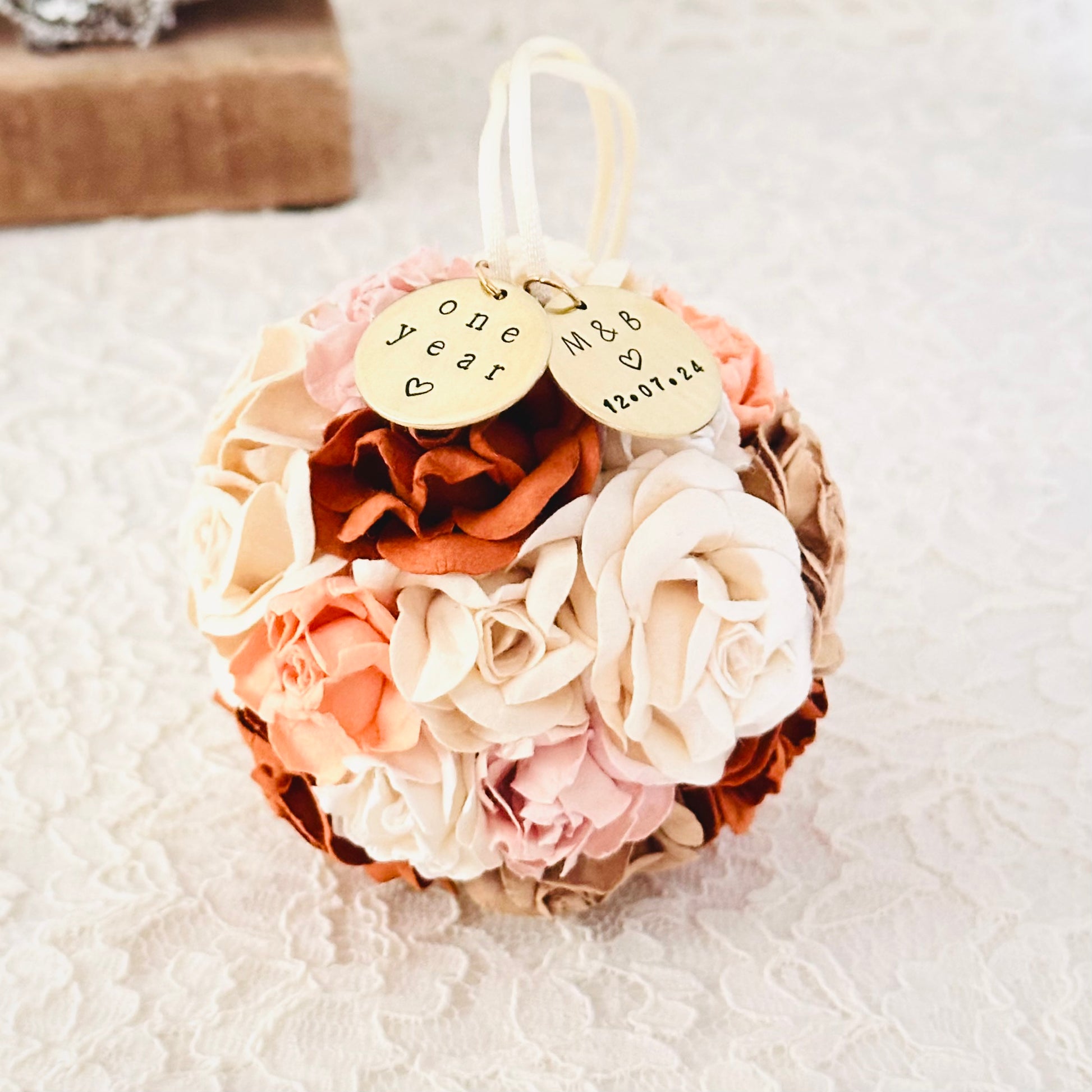 Decorative flower ornament with personalized charms on an ivory lace surface.  Flowers are in shades of terra cotta, peach, blush pink, ivory, and white and echo the wedding bouquet from the previous photo.