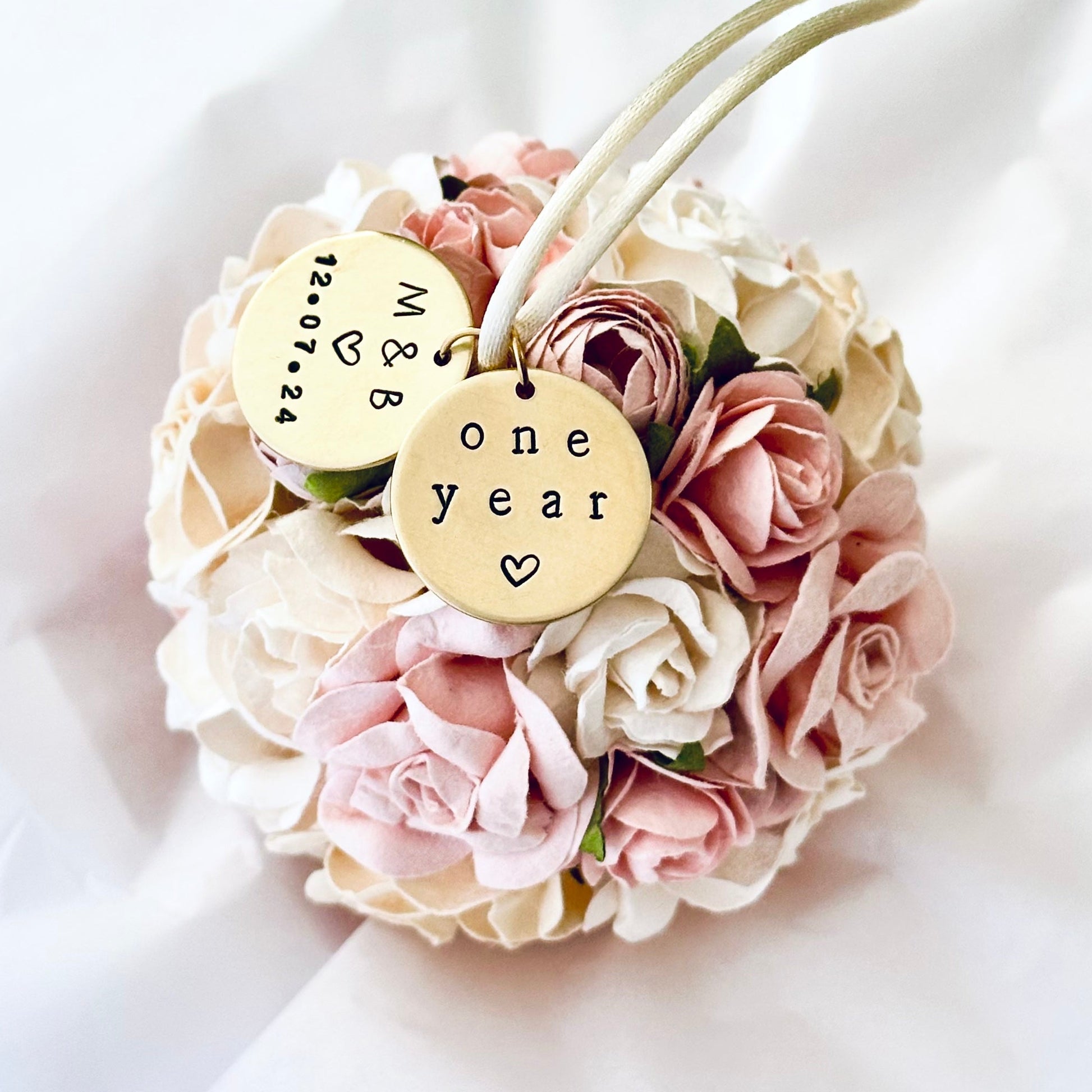 A round, ball-shaped flower ornament to celebrate the one year "paper" wedding anniversary.  The flowers are handmade of paper, and are detailed and elegant in shapes of roses and peonies.  The flowers are blush pink, white, ivory, and white.  The flower ornament rests on white tissue paper, and suggests a beautiful keepsake 1st anniversary gift.