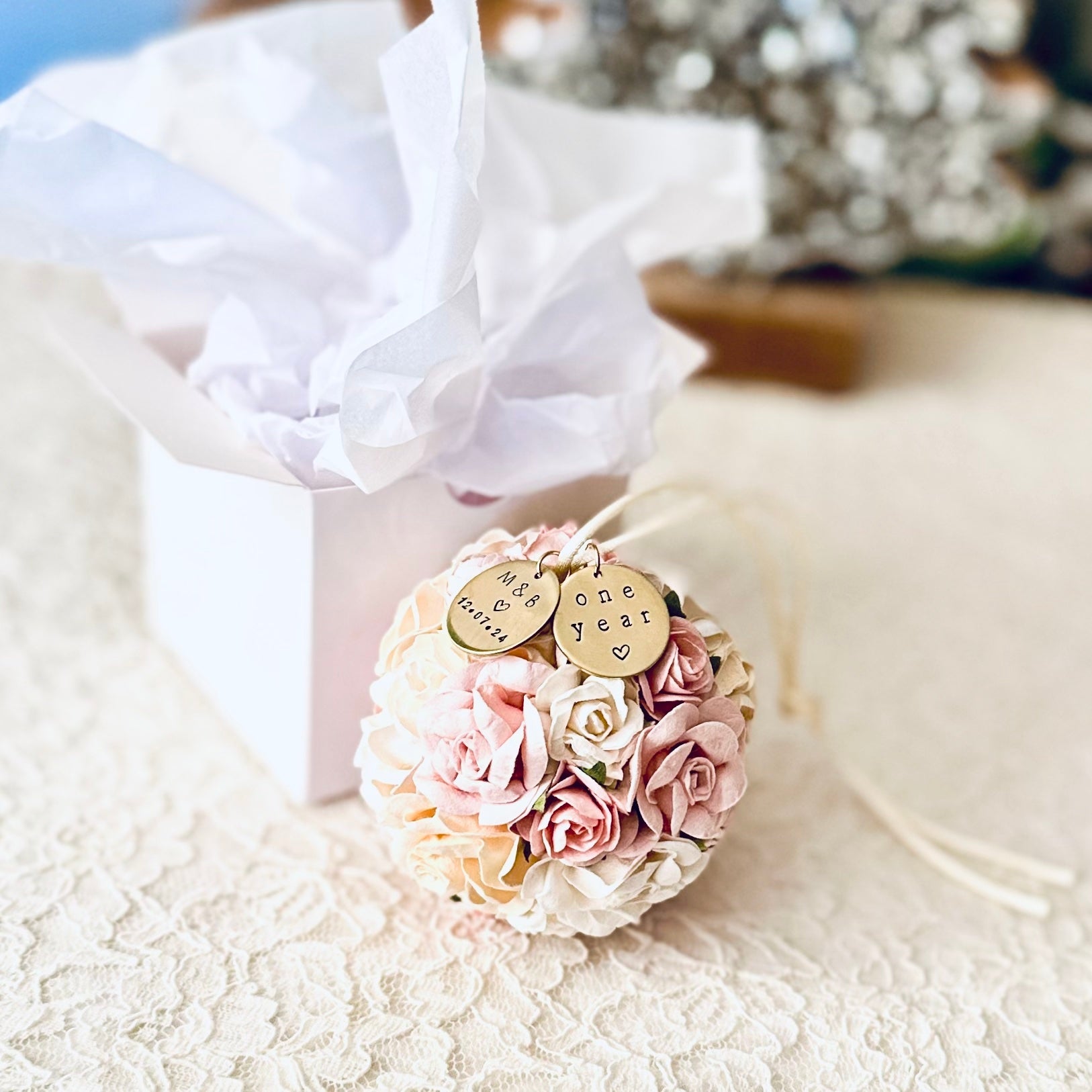 One Year Paper Flower anniversary ornament rests in front of a glossy white gift box.  The flowers are shades of blush pink, white, and ivory and are roses and peonies.
