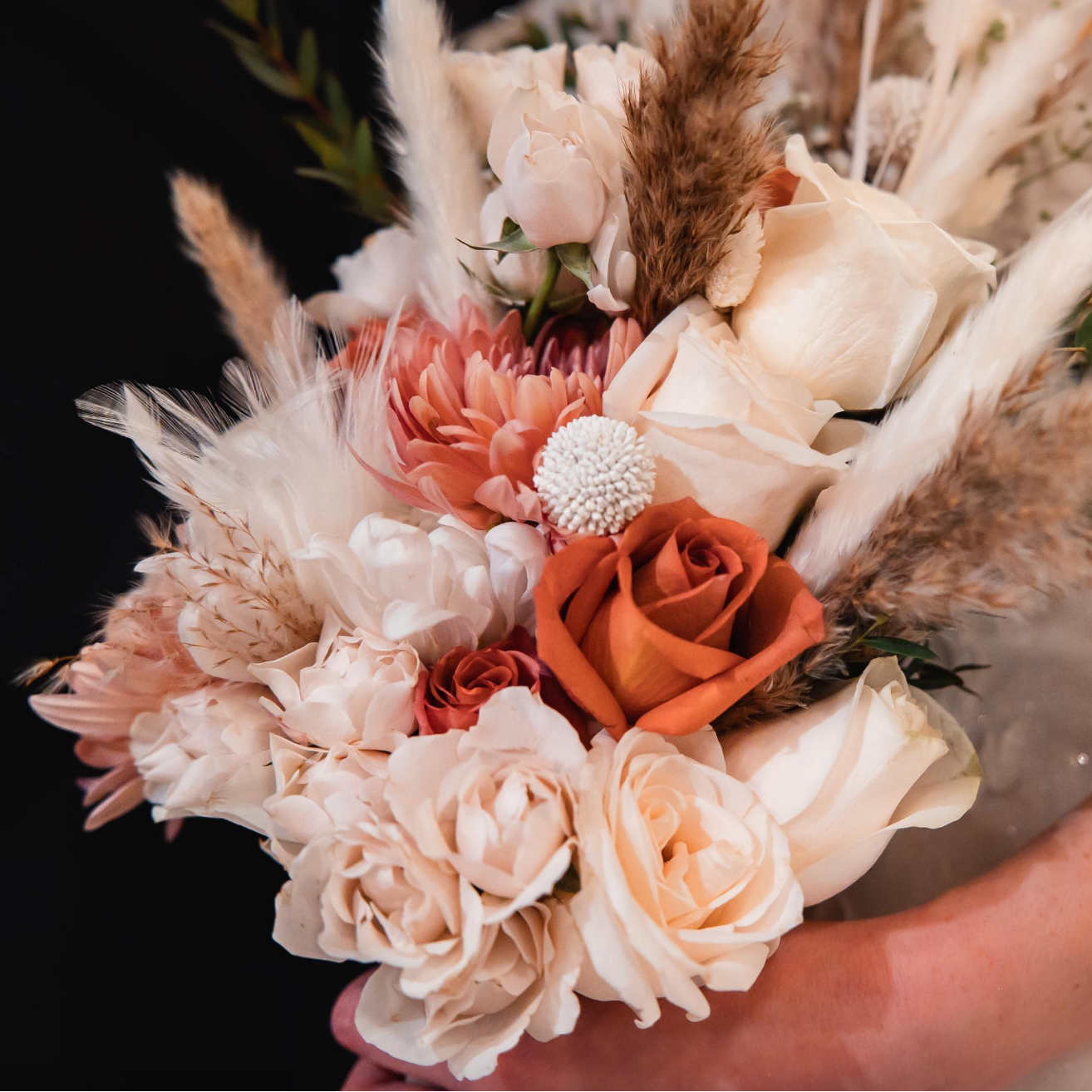 Wedding bouquet of flowers with roses in shades of terra cotta, ivory, blush pink, and peach held by a bride against a dark background.
