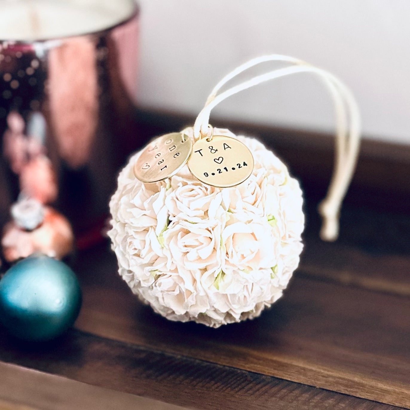 a one year paper flower anniversary ornament rests on a dark brown table.  It is made of white paper flowers, with 2 personalized gold colored charms attached.