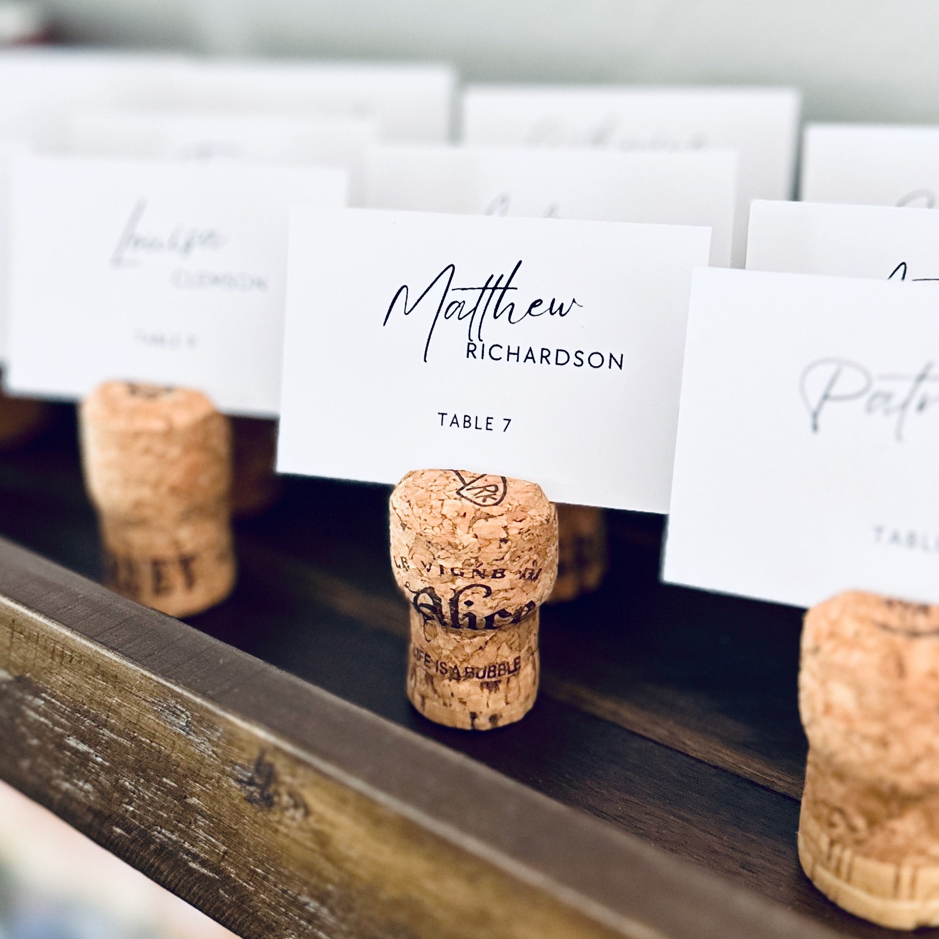 Champagne Cork place card holders with printed guest name cards on a rustic wooden shelf.