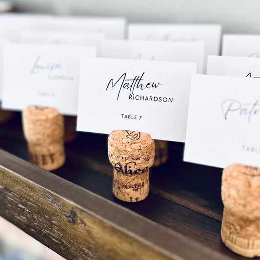 Champagne Cork place card holders with printed guest name cards on a rustic wooden shelf.