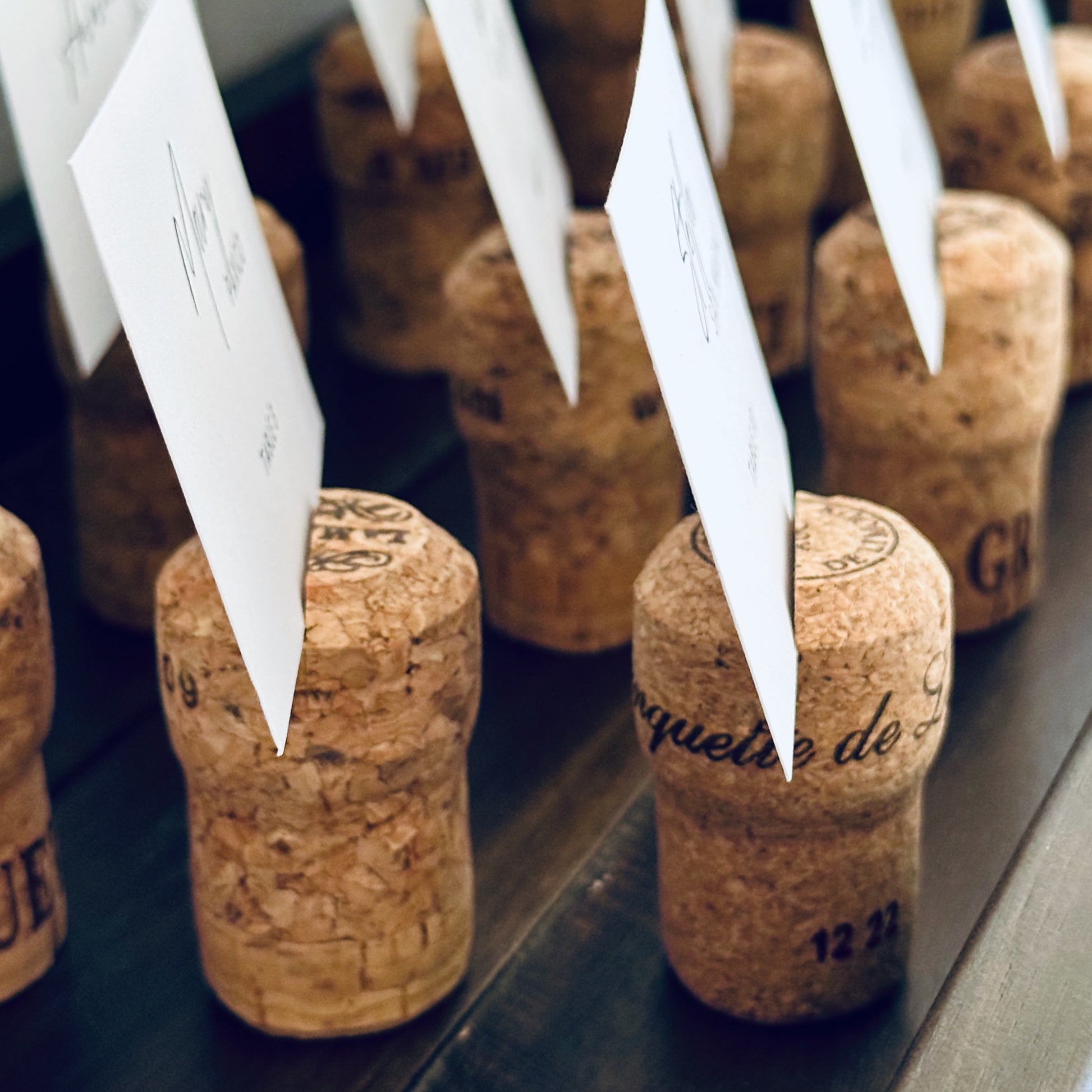 Champagne cork place card holders with white guest name cards displayed, used for wedding table assignments.  Corks and names are set up on a rustic wooden shelf.