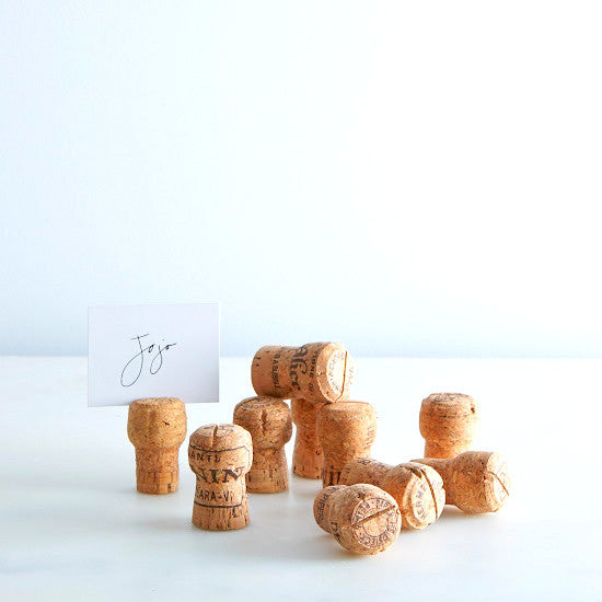 Several champagne cork place card holders displayed on a clean white surface with a card holder in the foreground showing a printed name card.